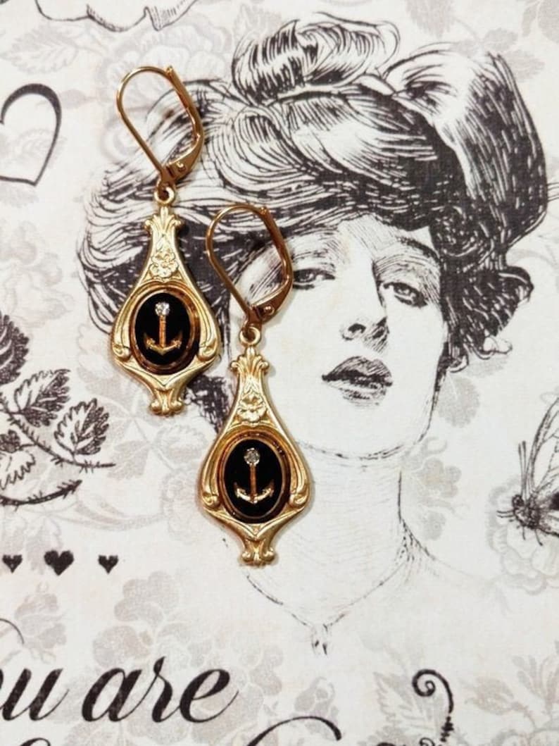 May include: Gold tone earrings with black enamel and a small anchor charm. The earrings have a teardrop shape and are accented with a clear rhinestone.