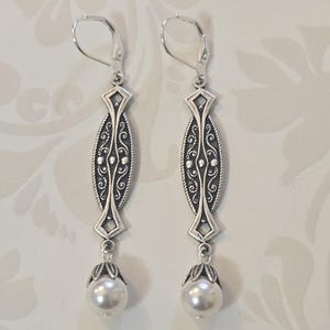 May include: A pair of silver-toned drop earrings. Each earring features an elongated, ornate design with a pearl at the bottom. The earrings have a lever-back closure. The design includes intricate scrollwork and geometric shapes.