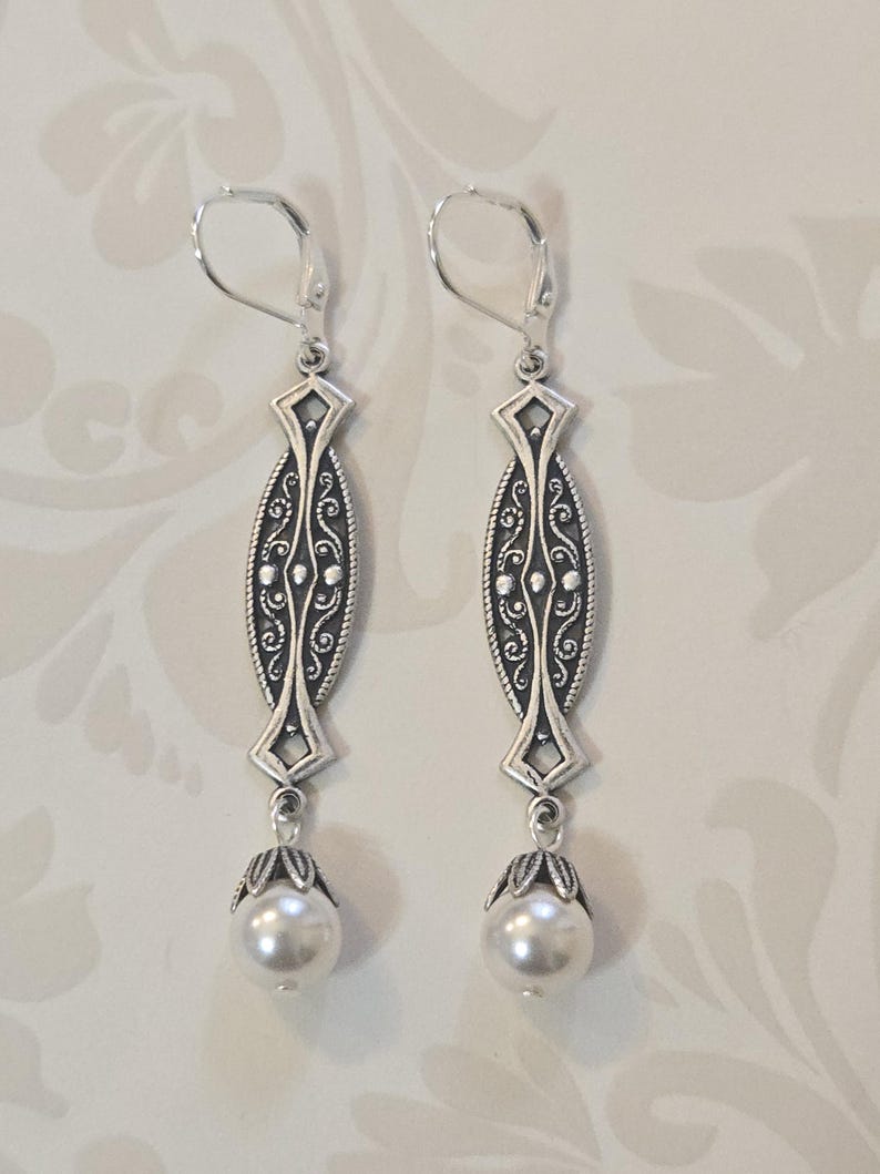 May include: A pair of silver-toned drop earrings. Each earring features an elongated, ornate design with a pearl at the bottom. The earrings have a lever-back closure. The design includes intricate scrollwork and geometric shapes.