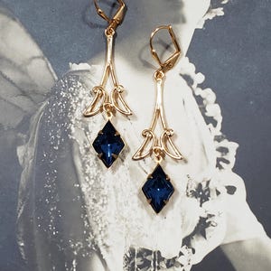 May include: A pair of gold dangle earrings with blue gemstone accents. The earrings feature a delicate, ornate design with a teardrop-shaped gemstone at the bottom.