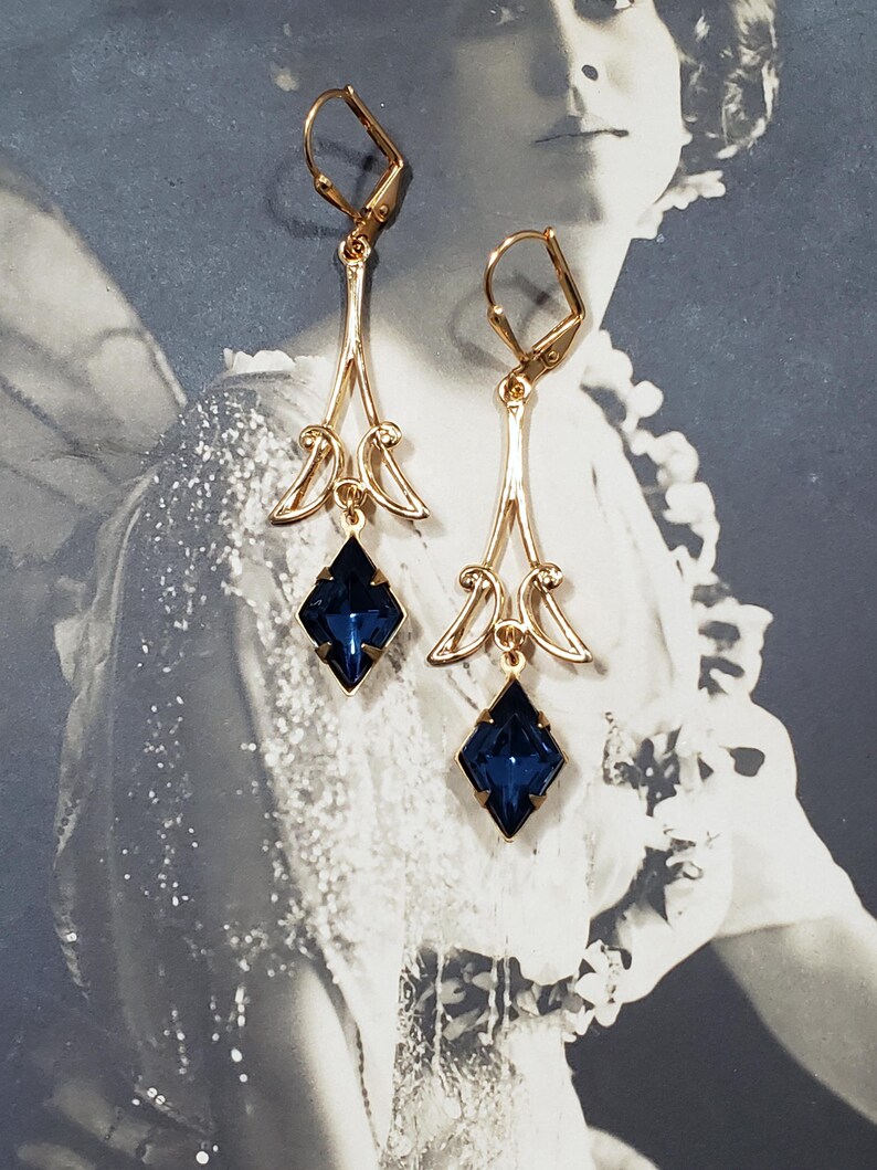 May include: A pair of gold dangle earrings with blue gemstone accents. The earrings feature a delicate, ornate design with a teardrop-shaped gemstone at the bottom.