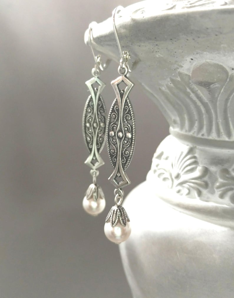 May include: Elegant silver-toned earrings with an ornate filigree design. Each earring features an elongated oval shape with intricate detailing and a teardrop-shaped pearl pendant. The earrings are suspended from a simple hook.