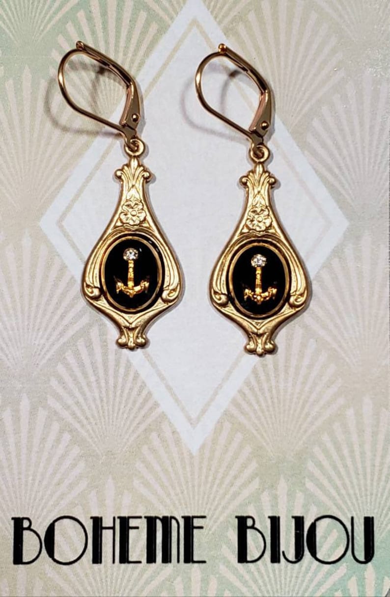May include: Gold dangle earrings with black enamel and a gold anchor charm. The earrings have a floral design and a small clear rhinestone on the anchor.