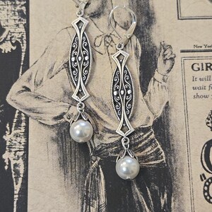 May include: A pair of silver-toned drop earrings. Each earring features an elongated, ornate central design with scrollwork and a diamond-shaped top. A faux pearl hangs from the bottom of each earring. The earrings are displayed against a vintage-style background.