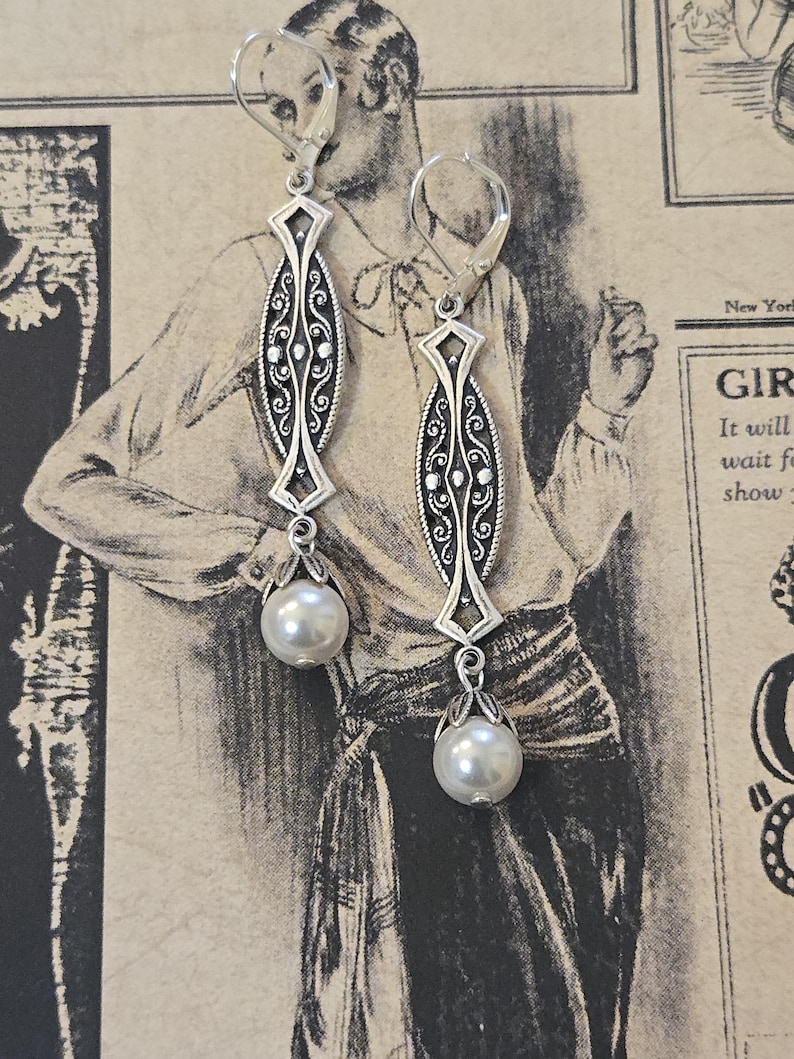 May include: A pair of silver-toned drop earrings. Each earring features an elongated, ornate central design with scrollwork and a diamond-shaped top. A faux pearl hangs from the bottom of each earring. The earrings are displayed against a vintage-style background.