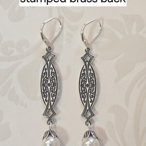 May include: A pair of silver-toned earrings with an ornate filigree design. Each earring features a long, oval-shaped pendant with intricate patterns and a small, round pearl at the bottom. The text "stamped brass back" is visible.