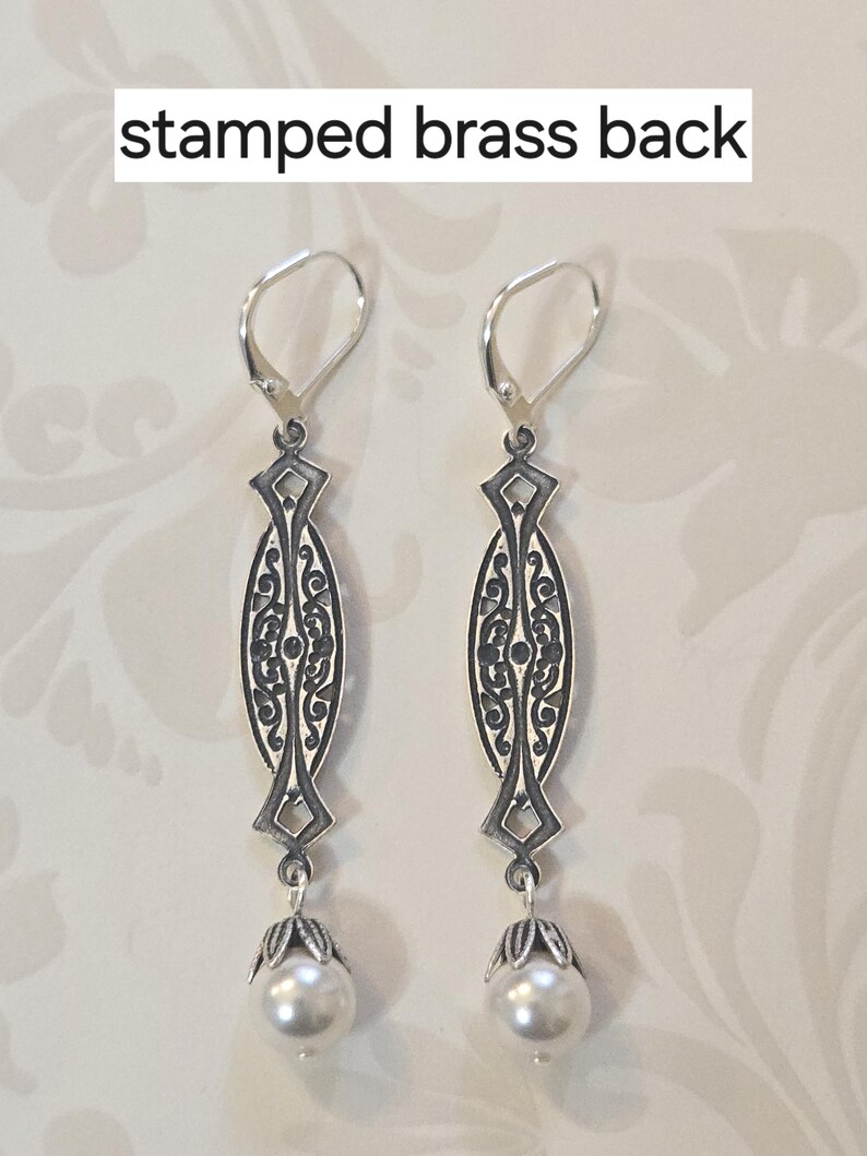 May include: A pair of silver-toned earrings with an ornate filigree design. Each earring features a long, oval-shaped pendant with intricate patterns and a small, round pearl at the bottom. The text "stamped brass back" is visible.