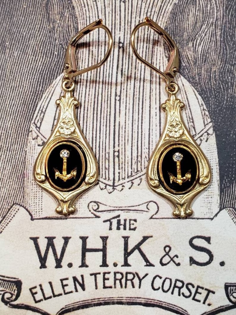 May include: Gold dangle earrings with black enamel and a gold anchor charm. The anchor is set with a small clear rhinestone.