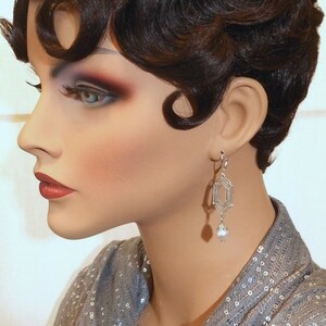 May include: A silver geometric dangle earring with a pearl drop. The earring is worn by a mannequin with short, dark brown hair styled in a 1920s bob.