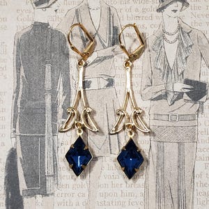 May include: A pair of gold dangle earrings with blue gemstone accents. The earrings feature a delicate, ornate design with a teardrop shape and a small, square-shaped gemstone at the bottom.