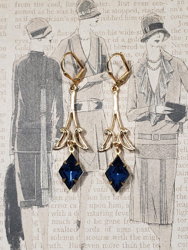 May include: A pair of gold dangle earrings with blue gemstone accents. The earrings feature a delicate, ornate design with a teardrop shape and a small, square-shaped gemstone at the bottom.