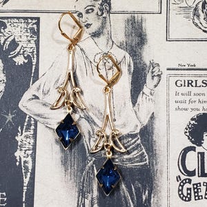 May include: A pair of gold dangle earrings with blue gemstone accents. The earrings feature a delicate, ornate design with a diamond-shaped gemstone at the bottom.