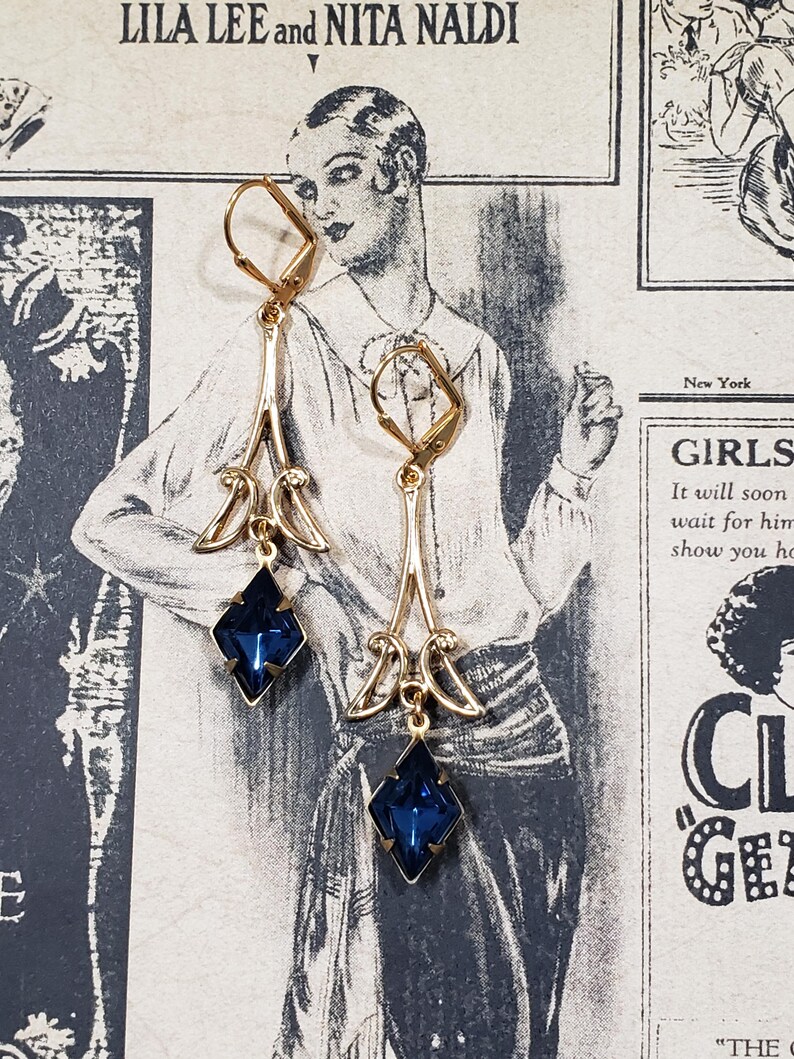 May include: A pair of gold dangle earrings with blue gemstone accents. The earrings feature a delicate, ornate design with a diamond-shaped gemstone at the bottom.