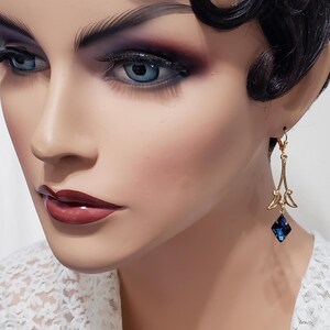 May include: Gold dangle earrings with a blue crystal teardrop. The earrings feature a delicate, intricate design.
