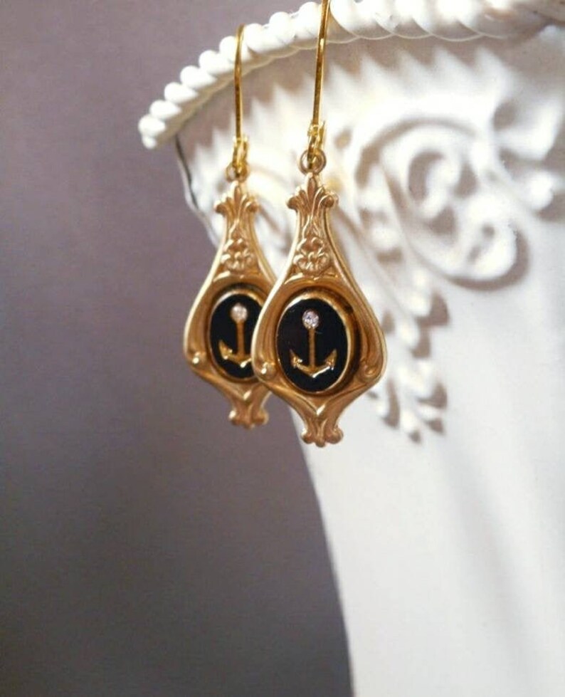 May include: Gold dangle earrings with a black enamel anchor charm and a single rhinestone. The anchor is set in a gold oval frame.
