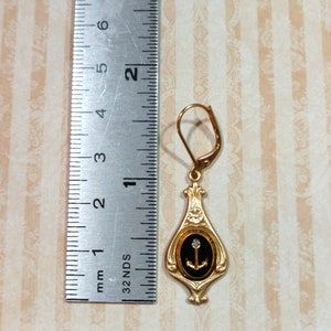 May include: Gold dangle earring with a black oval pendant featuring a gold anchor and a single clear rhinestone. The earring is approximately 1 inch long.