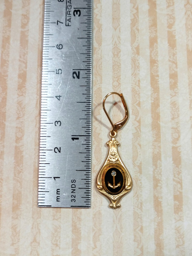 May include: Gold dangle earring with a black oval pendant featuring a gold anchor and a single clear rhinestone. The earring is approximately 1 inch long.