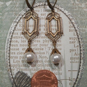 May include: A pair of gold-toned earrings with a geometric design and white pearl accents. The earrings are hanging from a penny.