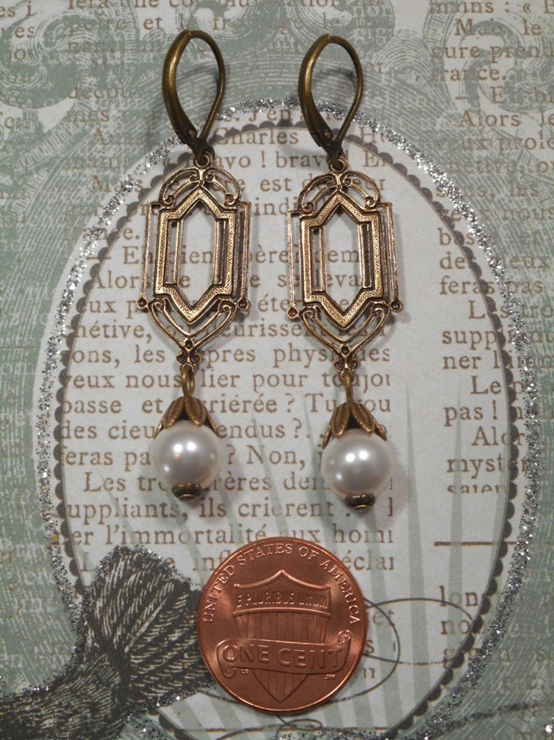May include: A pair of gold-toned earrings with a geometric design and white pearl accents. The earrings are hanging from a penny.