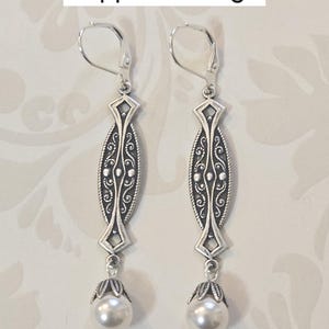 May include: Silver-toned flapper earrings with a decorative oval design and a dangling faux pearl. The earrings have a lever-back closure. The text "Flapper Earrings" is at the top of the image.