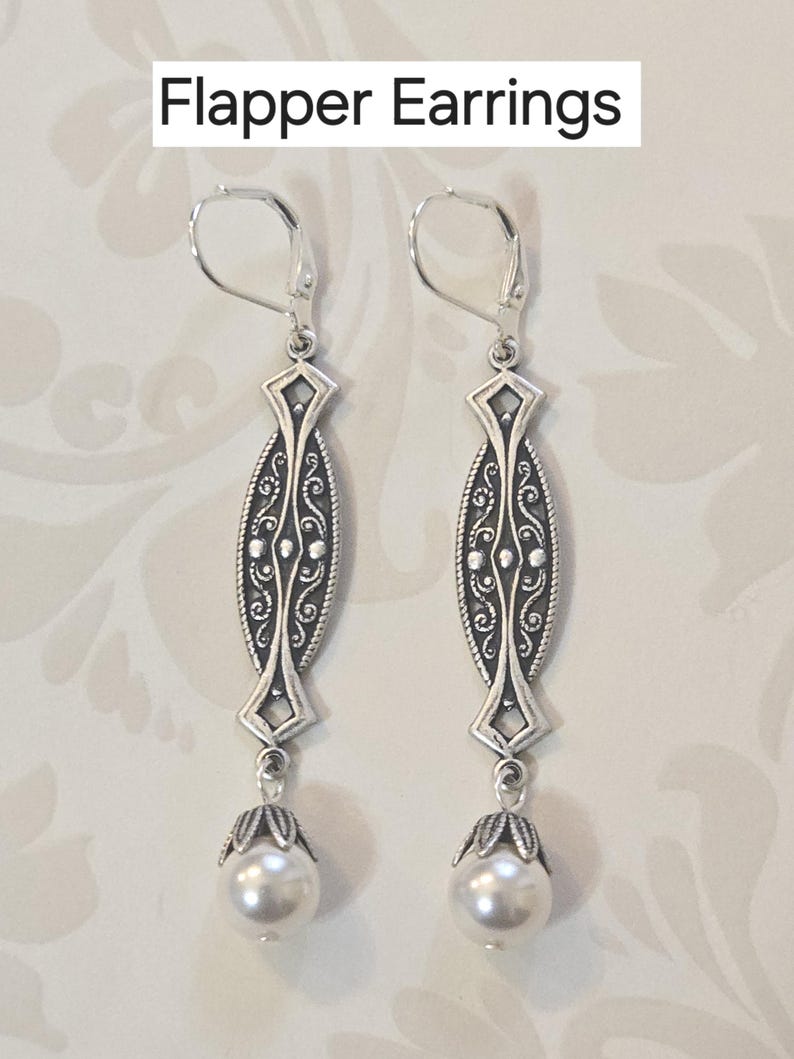 May include: Silver-toned flapper earrings with a decorative oval design and a dangling faux pearl. The earrings have a lever-back closure. The text "Flapper Earrings" is at the top of the image.