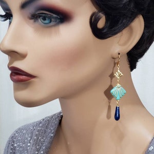 May include: A drop earring featuring a gold-tone metal fan-shaped top piece, a central turquoise-coloured diamond-shaped component with gold-tone ridges, and a smooth, dark blue teardrop-shaped bead dangling at the bottom of the earring structure.