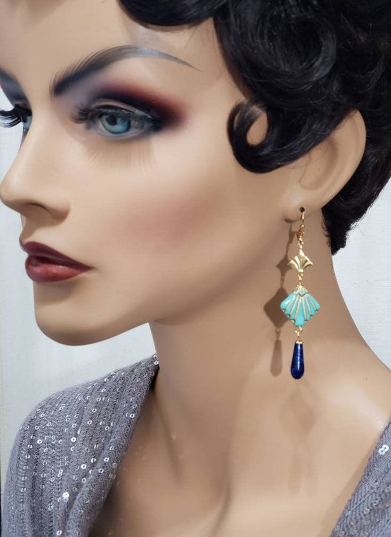 May include: A drop earring featuring a gold-tone metal fan-shaped top piece, a central turquoise-coloured diamond-shaped component with gold-tone ridges, and a smooth, dark blue teardrop-shaped bead dangling at the bottom of the earring structure.
