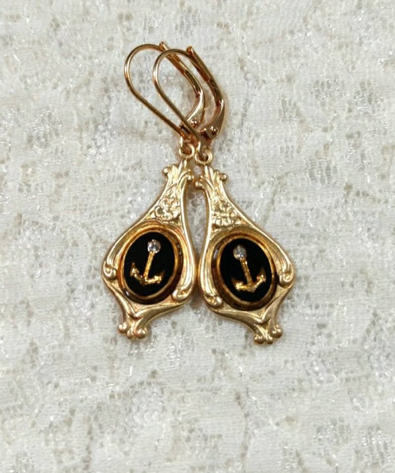 May include: Gold tone earrings with black enamel and a gold anchor design. Each earring has a clear rhinestone in the center of the anchor.