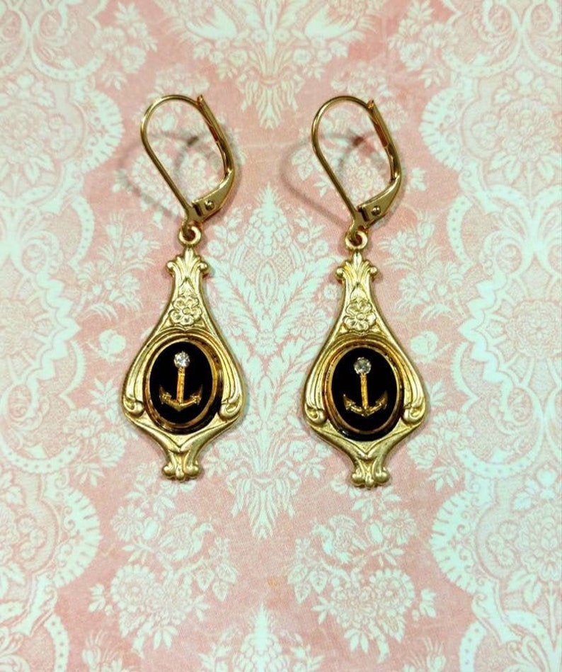May include: Gold dangle earrings with black enamel and a gold anchor with a single rhinestone in the center. The earrings have a teardrop shape with a decorative design.