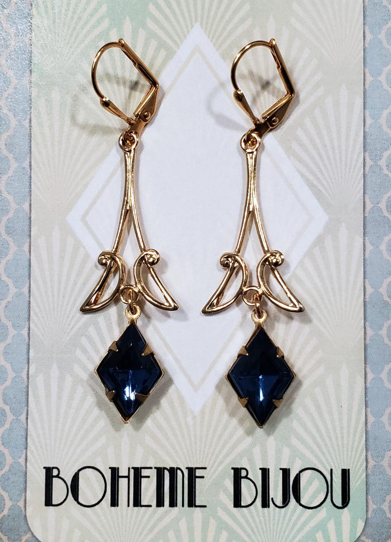 May include: A pair of gold dangle earrings with a dark blue gemstone pendant. The earrings feature a delicate, Art Deco-inspired design. The brand name "BOHEME BIJOU" is printed on the card.