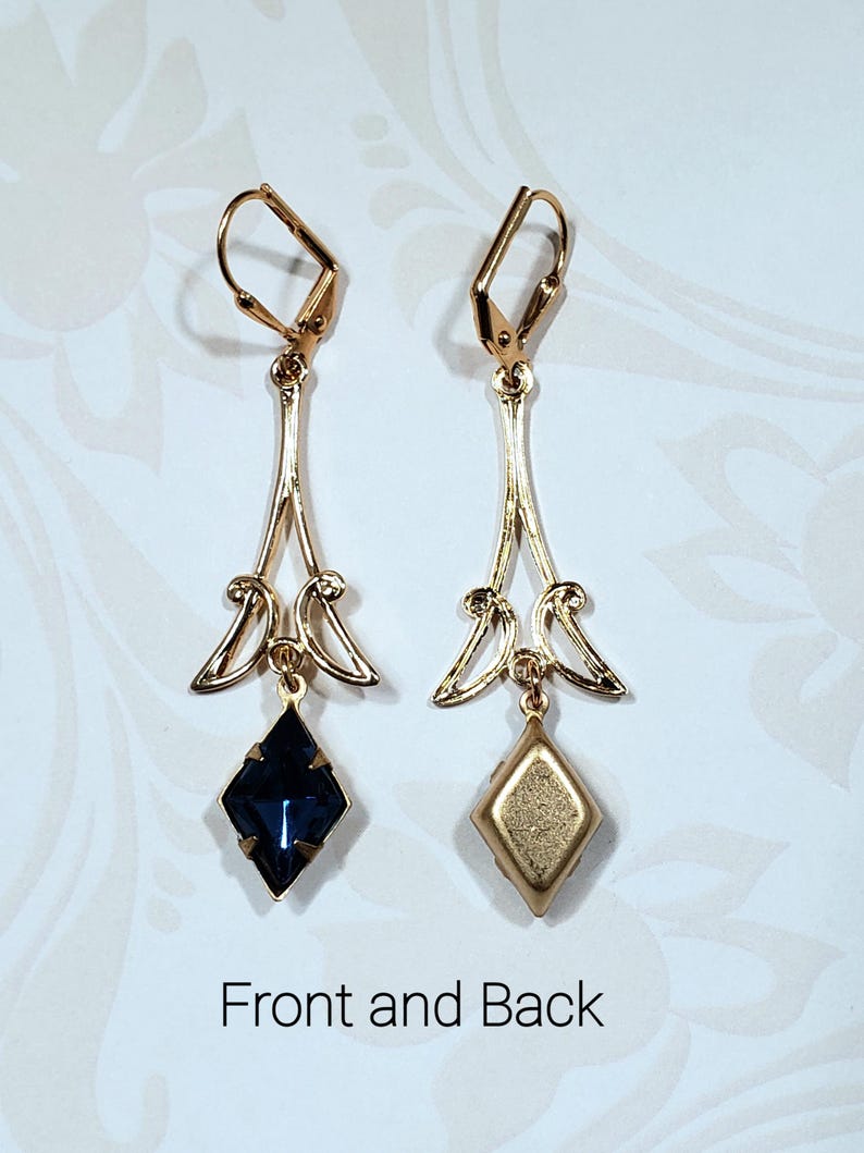 May include: A pair of gold dangle earrings with a dark blue faceted gemstone on one side and a gold back on the other. The earrings have a delicate, ornate design.
