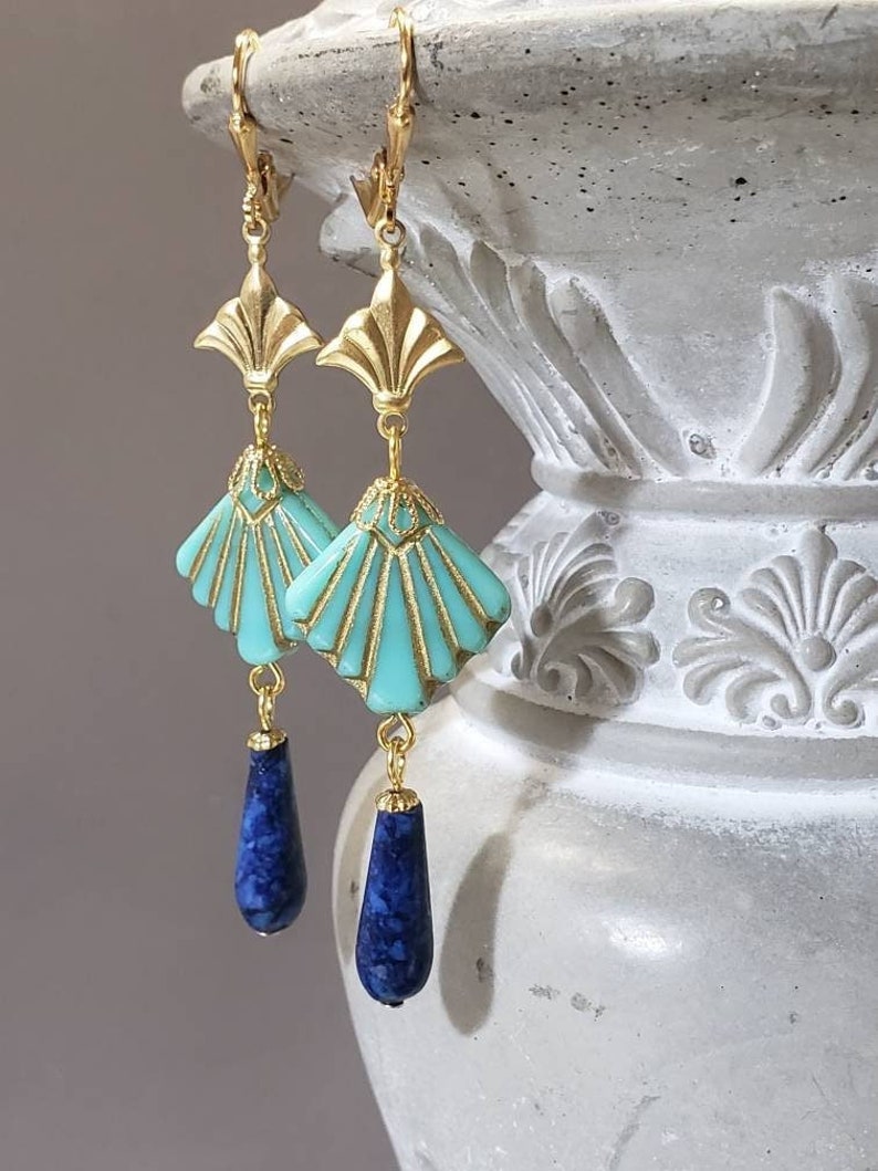 May include: A pair of gold-tone drop earrings featuring a fan-shaped turquoise component with gold detailing, suspended from a gold-tone metal topper. A dark blue, teardrop-shaped stone hangs from the base of each turquoise fan piece.