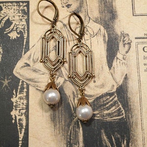 May include: A pair of gold-tone earrings with a geometric design and a pearl drop. The earrings have a filigree pattern and are suspended from a hook.