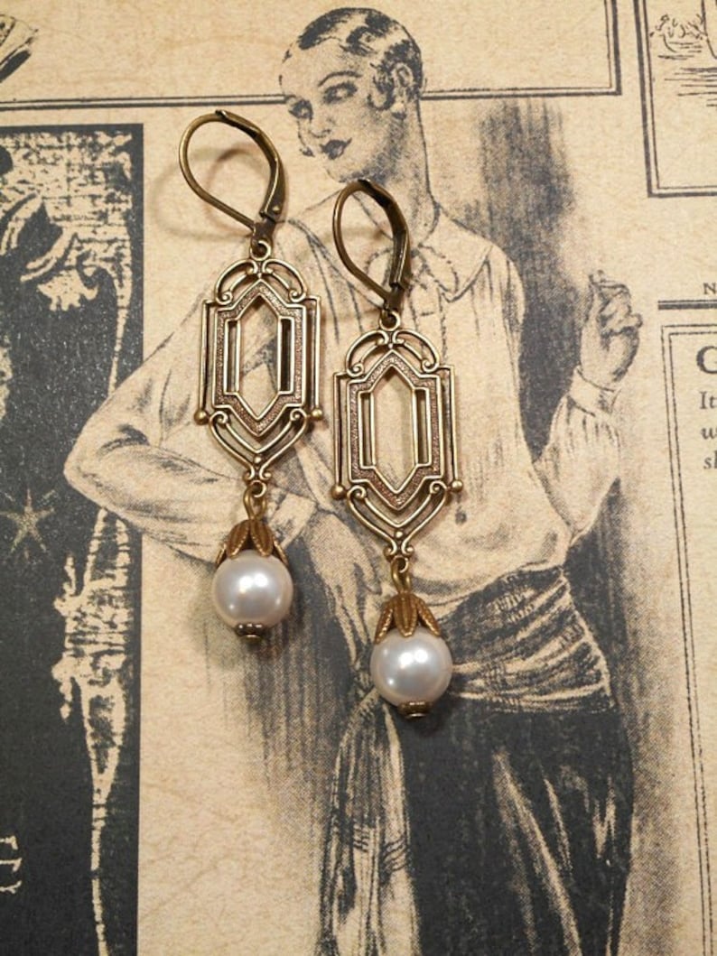May include: A pair of gold-tone earrings with a geometric design and a pearl drop. The earrings have a filigree pattern and are suspended from a hook.