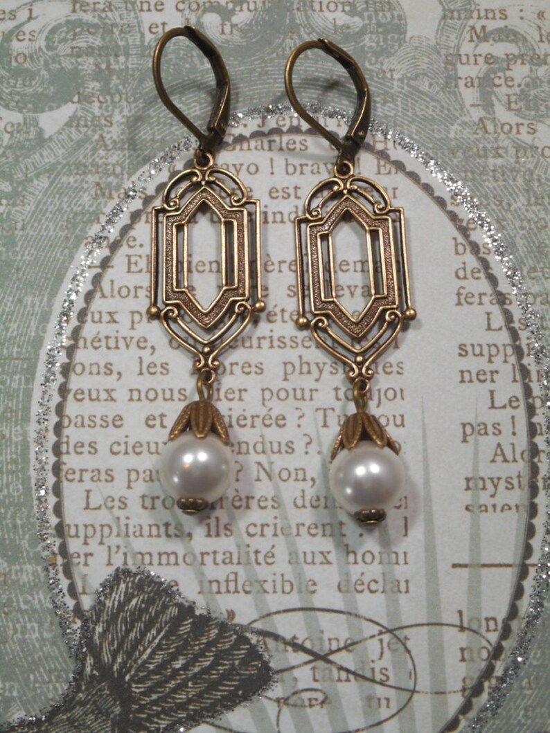 May include: A pair of gold-toned earrings with a geometric design and pearl accents. The earrings have a hook closure and are suspended from a delicate chain.