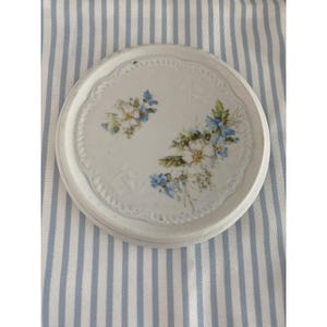 May include: A round white ceramic coaster featuring a delicate embossed scalloped edge and a central floral motif. The design displays small clusters of white and blue flowers with green leaves painted in a soft, detailed watercolor style.
