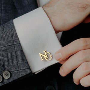 May include: Gold-colored cufflinks with the initials "NG" are displayed on a white dress shirt cuff. The cuff is part of a formal outfit, including a gray plaid suit jacket. The person is adjusting the cufflink.