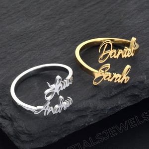 Può includere: Due anelli personalizzati con nomi, uno argentato e uno dorato, esposti su una superficie scura. L'anello argentato presenta i nomi "Laura" e "Andrew" in corsivo. L'anello dorato mostra i nomi "Daniel" e "Sarah".