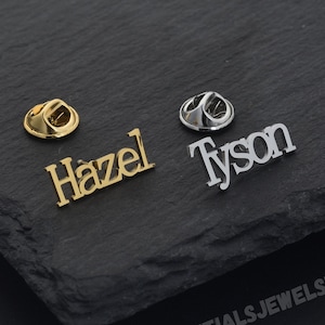 Op de afbeelding: Twee gepersonaliseerde naamspelden, één goudkleurig met de naam "Hazel" en één zilverkleurig met de naam "Tyson". De spelden worden gepresenteerd op een donker, getextureerd oppervlak. De achterkanten van de spelden zijn ook zichtbaar.
