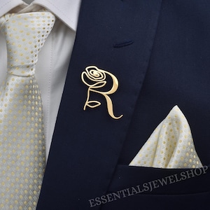 Può includere: Spilla da bavero a forma di rosa dorata su una giacca da abito blu navy. La spilla ha la forma di una rosa stilizzata con la lettera "R" incorporata. Sono visibili anche una cravatta e un fazzoletto da taschino con motivi bianchi e dorati.