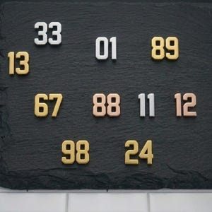 Pode incluir: Uma coleção de enfeites metálicos em forma de número em várias cores, incluindo ouro, prata e ouro rosa. Os números estão dispostos sobre uma superfície cinza escuro. Os números incluem 01, 11, 12, 13, 24, 33, 67, 88, 89 e 98.
