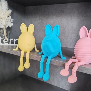 Könnte beinhalten: Drei bunte Hasenfiguren mit langen Beinen und Ohren. Der Hase links ist gelb, der mittlere Hase ist blau und der Hase rechts ist rosa.