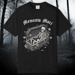Puede incluir: Camiseta negra con una ilustración detallada de un esqueleto en un ataúd, rodeado de vides y flores. Las palabras "Memento Mori" están impresas encima de la ilustración en una fuente adornada. La camiseta está hecha de un material suave.