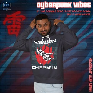 Op de afbeelding: Donkergrijze hoodie met de tekst "SAMURAI CHIPPIN' IN" en een rode afbeelding van een hand die een apparaat vasthoudt. De hoodie heeft ook de tekst "cyberpunk vibes" en "IF YOUR DEFAULT MODE IS NOT BACKING DOWN, THIS IS YOUR HOODIE."
