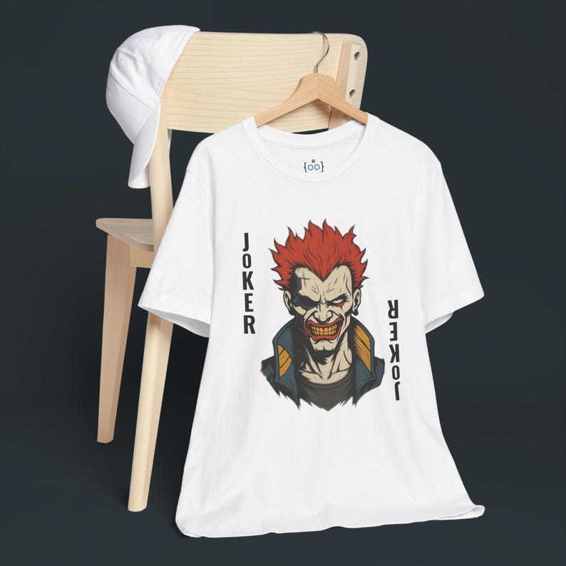 Peut inclure: T-shirt blanc avec un dessin du Joker aux cheveux roux et un sourire sinistre. Le mot "JOKER" est imprim&eacute; verticalement des deux c&ocirc;t&eacute;s du dessin. Une casquette de baseball blanche est pos&eacute;e sur une chaise en bois.