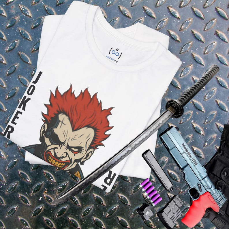 Peut inclure: T-shirt blanc avec un motif du Joker et le mot "JOKER" en noir. Un katana, un pistolet jouet bleu et rouge et d'autres accessoires sont dispos&eacute;s sur une surface m&eacute;tallique &agrave; motifs de losanges. Le t-shirt porte le texte "{oo}" et "GEEKIFIED".