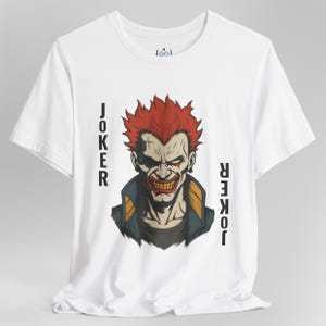 Peut inclure: T-shirt blanc avec un motif du Joker aux cheveux rouges et un sourire mena&ccedil;ant. Le mot "JOKER" est imprim&eacute; verticalement de chaque c&ocirc;t&eacute; de l'image. Le t-shirt a un col rond et des manches courtes.