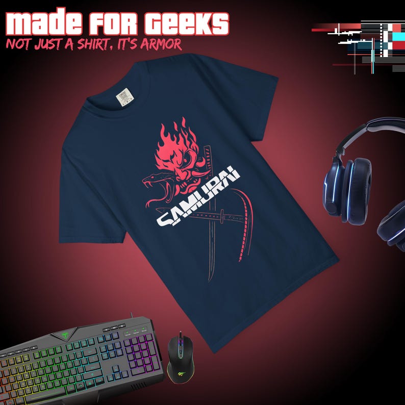 May include: Navy blue t-shirt with a red and white graphic of a samurai mask, swords, and the word "SAMURAI". The text "MADE FOR GEEKS" and "NOT JUST A SHIRT, IT'S ARMOR" are at the top. A gaming keyboard, mouse, and headphones are also visible.