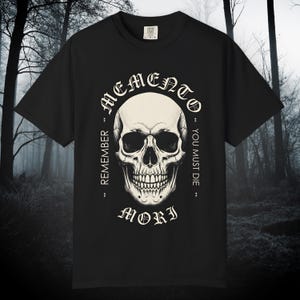 Puede incluir: Camiseta negra con un gráfico detallado de calavera y las palabras "Memento Mori" en un diseño circular. El diseño incluye las frases "Remember" y "You Must Die". La camiseta tiene cuello redondo y mangas cortas.
