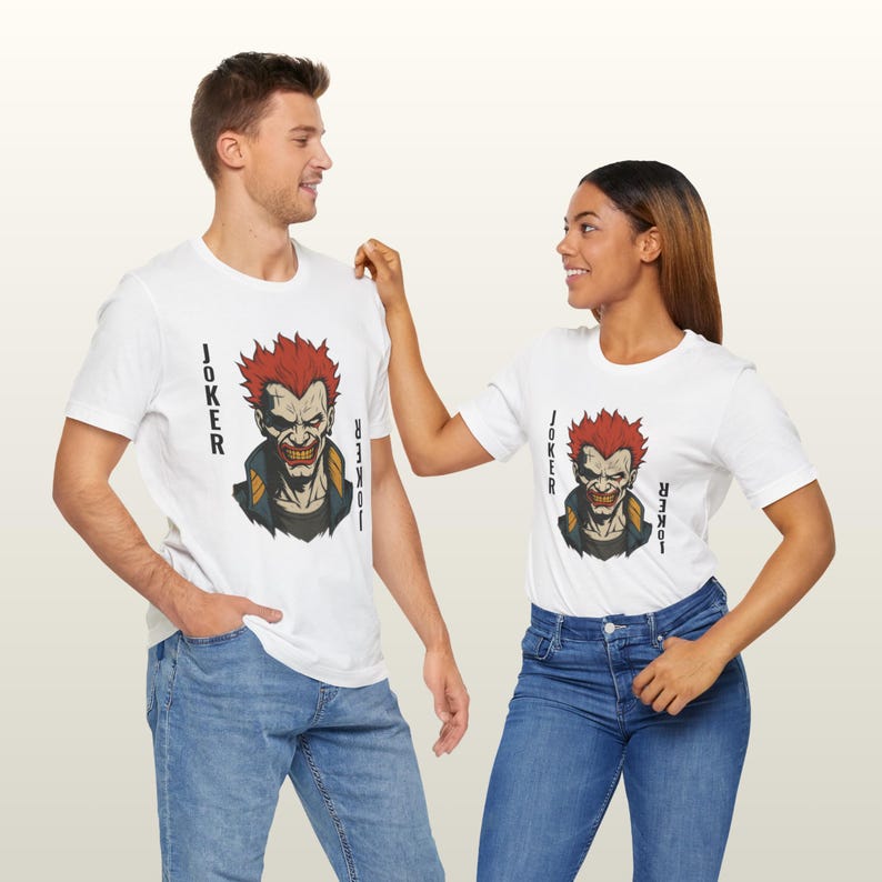 Peut inclure: T-shirts blancs avec un graphisme du Joker aux cheveux roux et un sourire sinistre. Le mot "JOKER" est imprim&eacute; verticalement sur les c&ocirc;t&eacute;s. Les mod&egrave;les portent des jeans bleus.
